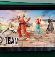 A handheld gaming console displays a screenshot of a fighting game featuring cartoonish characters in dynamic poses. The characters are standing in a virtual arena setting with a sky and cloud backdrop. The console is placed on a green surface.